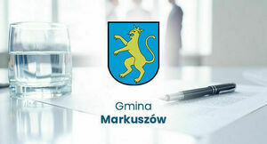 Na zdjęciu widoczny jest herb Gminy Markuszów przedstawiający żółtego lwa na niebieskim tle, umieszczony na biurku z papierami, długopisem i szklanką wody.