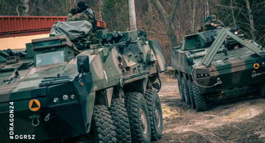 Pojazdy wojskowe i żołnierze w terenie leśnym. Na wozach oznaczonych symbolem Dragon z żółtym trójkątem są umundurowani żołnierze.