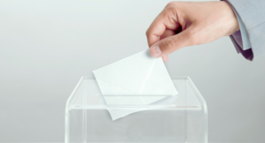 Osoba wkłada kartkę do głosowania do przezroczystej urny wyborczej na szarym tle.