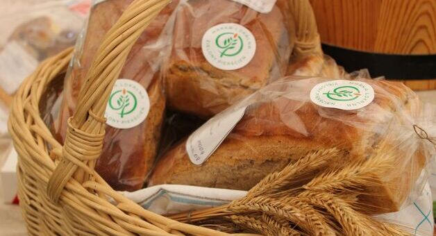 Zdjęcie przedstawiające pakowane chleby w przezroczystych foliowych opakowaniach z etykietami, umieszczone w plecionym koszyku obok kłosów zboża.