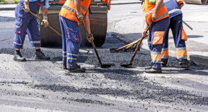 Pracownicy drogowi w odblaskowych kamizelkach naprawiają asfalt, używając łopat i grabi.