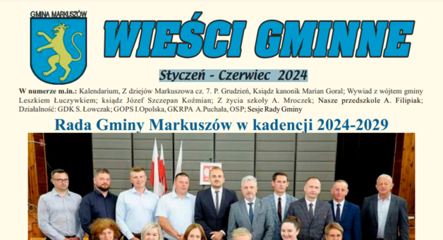 Grupa osób stoi i siedzi w dwóch rzędach, mężczyźni w garniturach i kobiety w eleganckich strojach, na tle flag i emblematu gminy.