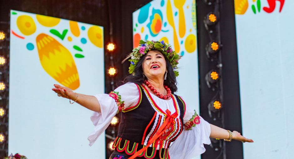 Kobieta w kolorowym stroju ludowym tańczy na scenie podczas festiwalu. Obok niej dwie inne kobiety w strojach ludowych. W tle dekoracje z motywami kuchennymi i logo festiwalu.