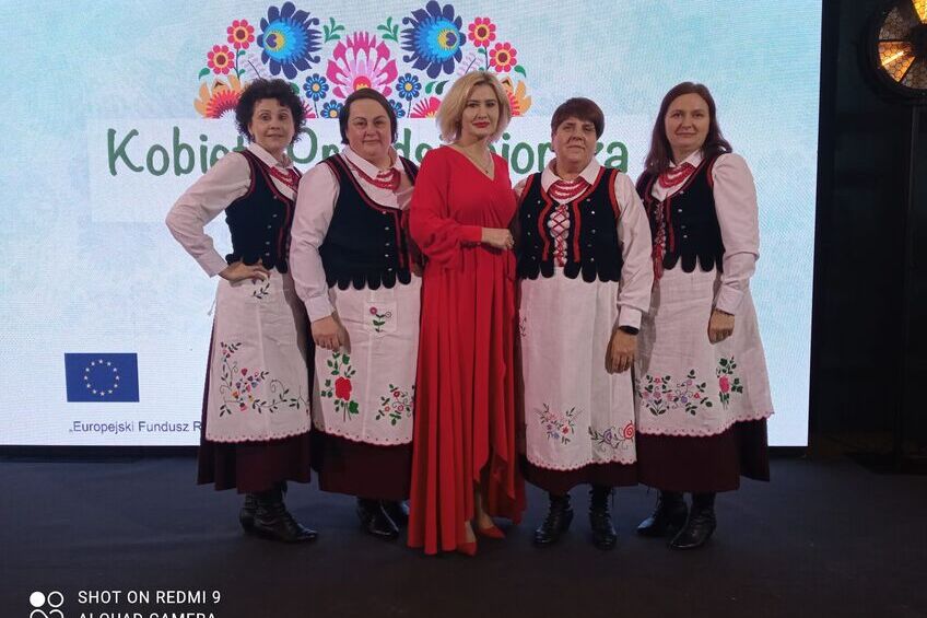 Grupa pięciu kobiet pozuje na scenie; jedna w czerwonej sukni, cztery w tradycyjnych strojach ludowych, w tle ścianka z kwiatowym wzorem i napisem "Kobiety Odpowiedzialne Lokalnie".