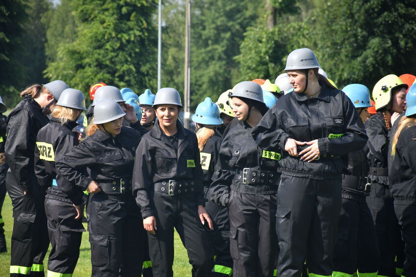 Grupa osób w czarnych uniformach i kaskach stojąca na trawiastym terenie.