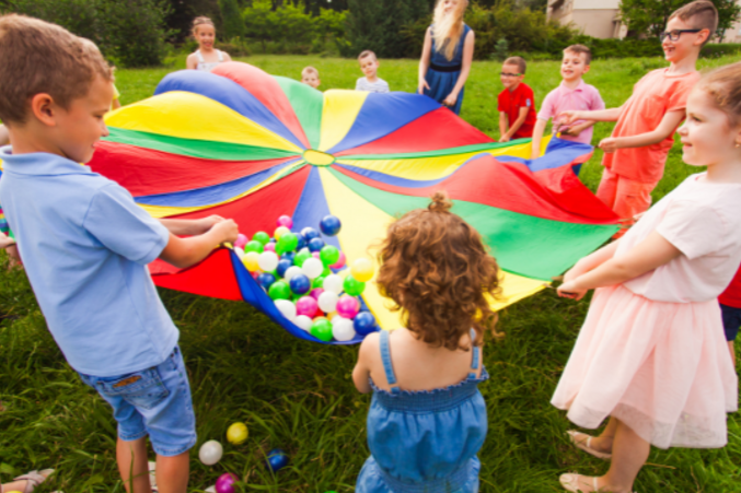 Dzieci bawią się kolorowym spadochronem z piłkami podczas zabawy na trawie, dorosły nadzoruje.