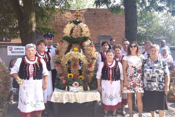 Grupa osób w tradycyjnych strojach ludowych prezentuje wielki, kolorowy wieniec dożynkowy, stoją przed historycznym budynkiem.