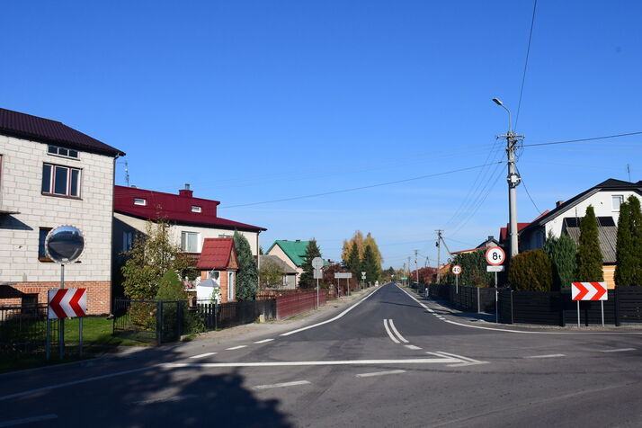 Zdjęcie przedstawia słoneczną, asfaltową drogę w małym mieście z domami po obu stronach i znakami drogowymi.