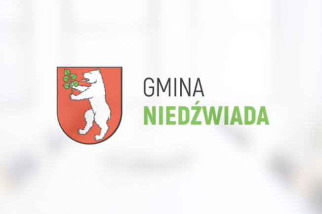 Herb Gminy Niedźwiada przedstawiający białego niedźwiedzia trzymającego zieloną gałązkę na czerwonym tle, powyżej napis "GMINA NIEDŹWIADA".