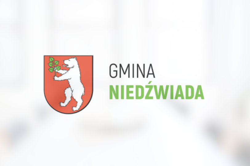 Herb Gminy Niedźwiada przedstawiający białego niedźwiedzia trzymającego zieloną gałązkę na czerwonym tle, powyżej napis "GMINA NIEDŹWIADA".