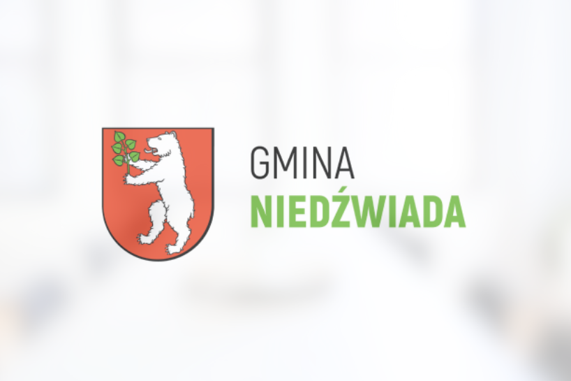 Herb Gminy Niedźwiada przedstawiający białego niedźwiedzia trzymającego zieloną gałązkę na czerwonym tle, powyżej napis "GMINA NIEDŹWIADA".