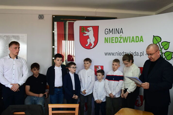 W sali na tle baneru z logo Gminy Niedźwiada stoi grupa młodych chłopców i kilku dorosłych mężczyzn. Jeden z dorosłych czyta dokument, reszta patrzy na niego.