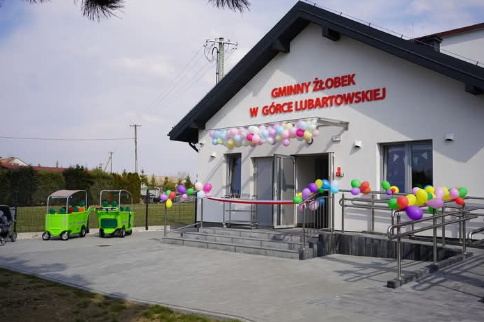 Budynk gminnego żłobka w Górce Lubartowskiej. Fasada ozdobiona balonami, widoczny wózek dziecięcy oraz miniaturowy wagonik. Przed wejściem znajdują się schody i podjazd dla wózków.