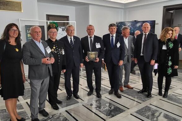 Grupa osób w eleganckich strojach pozuje do zdjęcia wewnątrz budynku. Niektórzy trzymają nagrody lub medale. Pomieszczenie ma marmurową podłogę i jasne ściany. W tle widoczne drzwi i plakat.