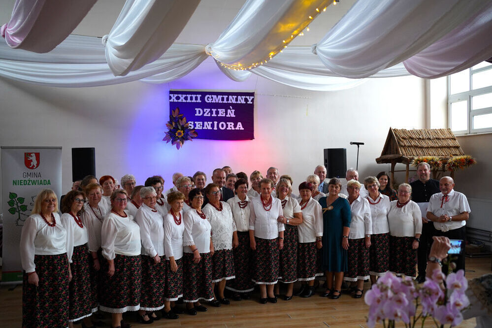 Grupa seniorów w białych bluzkach i ciemnych spódnicach pozuje wspólnie na scenie. Nad nimi baner "XXXIII Gminny Dzień Seniora". Sala udekorowana w białe materiałowe draperie i stoi tuż obok małej chatki modelowej.