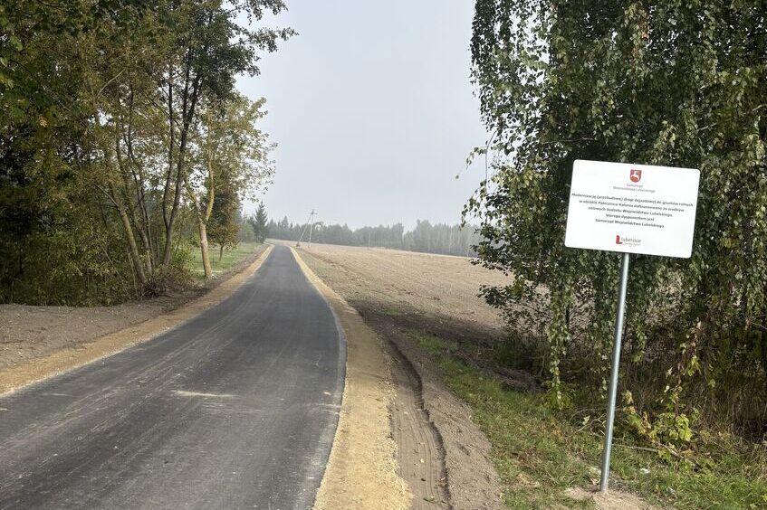 Wąska asfaltowa droga prowadząca przez wiejski krajobraz. Po lewej stronie widać drzewa, po prawej pola i znak informacyjny na skraju drogi. Niebo jest zachmurzone.