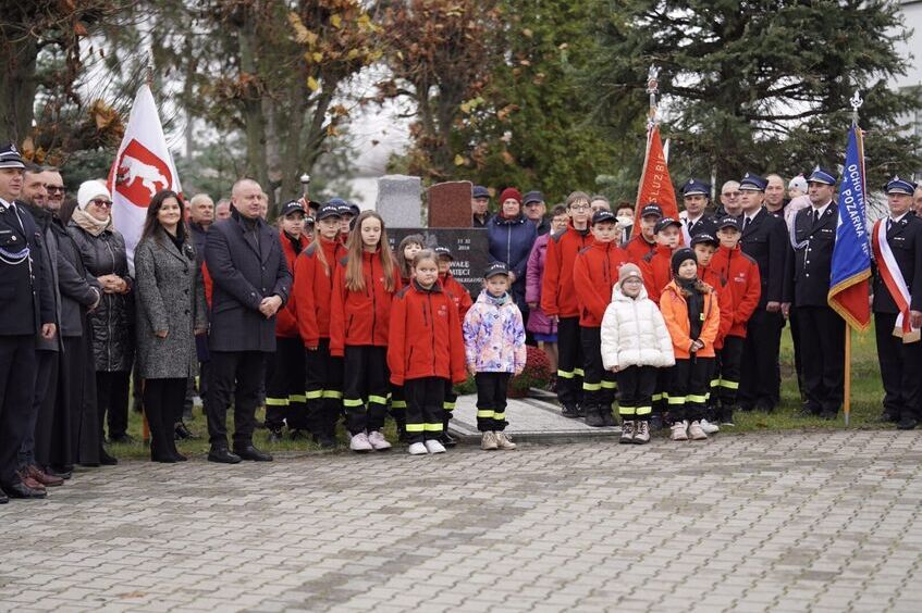 Grupa ludzi, w tym dzieci w czerwonych kurtkach, stoi obok pomnika na zewnątrz. Niektórzy trzymają flagi. W tle widać drzewa. Atmosfera wskazuje na oficjalne wydarzenie lub uroczystość.