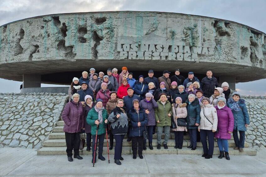 Grupa seniorów pozuje do zdjęcia przed pomnikiem z napisem "Los nasz dla was przestrogą". Tło stanowi kamienny mur i pochmurne niebo. Wszyscy są ciepło ubrani.
