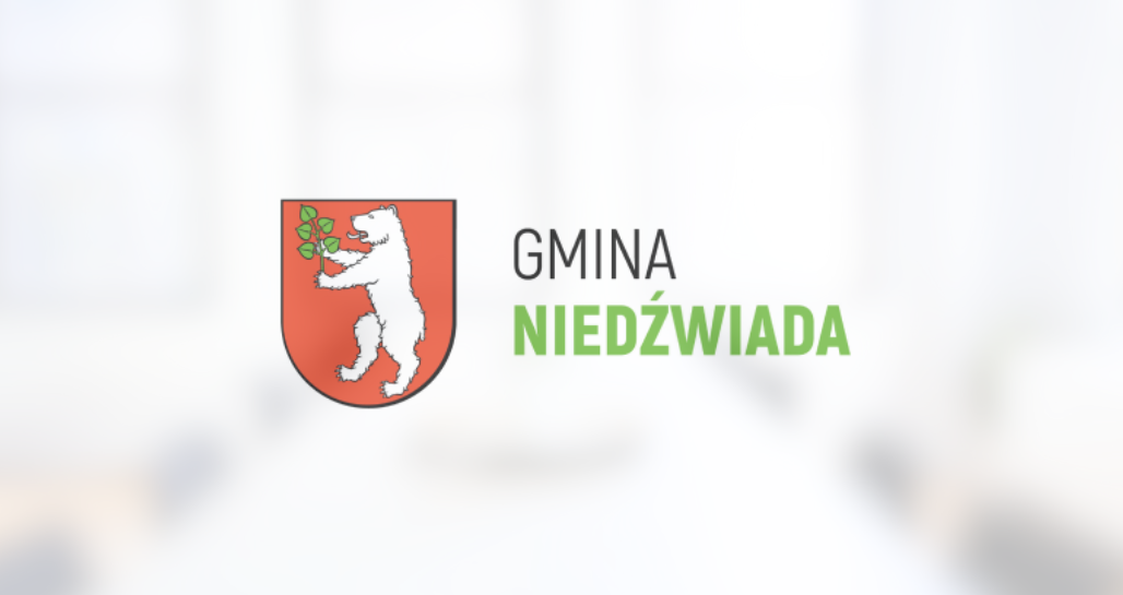 Herb Gminy Niedźwiada przedstawiający białego niedźwiedzia trzymającego zieloną gałązkę na czerwonym tle, powyżej napis "GMINA NIEDŹWIADA".