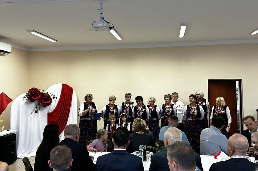 Grupa ludzi na scenie w odświętnych strojach wykonuje utwór. Przed nimi przy stole siedzą widzowie. W tle dekoracja z czerwonymi różami i biało-czerwonym materiałem.