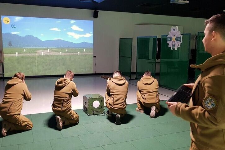 Na zdjęciu grupa czterech osób w mundurach trenuje strzelanie w symulatorze strzelnicy. Patrzą na projekcję przedstawiającą krajobraz. Jedna osoba stoi obok, trzymając kontroler lub notatnik.