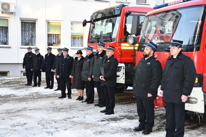 Grupa strażaków w czarnych mundurach stoi na zewnątrz przed dwoma czerwonymi wozami strażackimi. Tło stanowi biały budynek. Ziemia pokryta jest śniegiem.