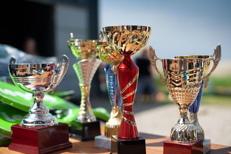 Na zdjęciu widać  pięć różnorodnych, błyszczących pucharów w złotym i srebrnym kolorze. Stoją na stole na zewnątrz przy słonecznym dniu. W tle rozmyta sceneria.