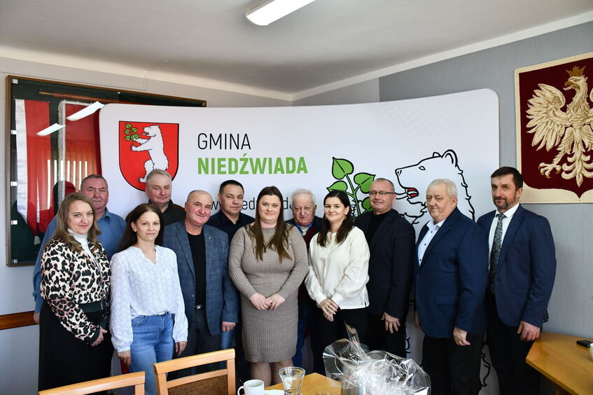 Grupa osób stoi w pomieszczeniu przed banerem z napisem "Gmina Niedźwiada". W tle widać herb z niedźwiedziem. Osoby pozują do zdjęcia, są uśmiechnięte.