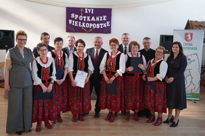 Grupa osób w tradycyjnych strojach ludowych pozuje obok kilku elegancko ubranych osób. W tle widać baner z napisem "XVI Spotkanie Wielkopostne" oraz logo gminy.