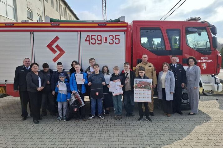 Grupa ludzi, w tym dzieci i dorośli, pozuje przed czerwonym wozem strażackim z boku budynku. Dzieci trzymają dyplomy i plakat. Pogoda jest pochmurna.