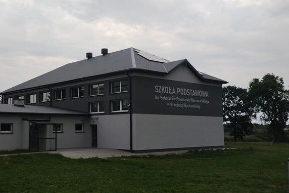 Nowoczesny budynek szkoły w odcieniach szarości, z napisem na ścianie. Otoczony jest zielonym trawnikiem, a w tle widać drzewa i pochmurne niebo.