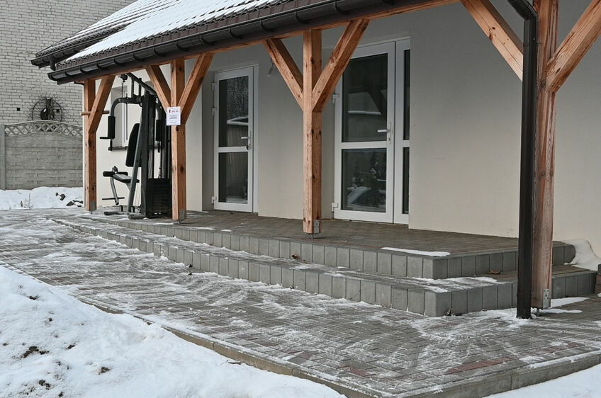 Zdjęcie przedstawia wejście do nowoczesnego budynku z drewnianymi elementami. Widoczne są schody i pochylnia dla wózków, wszystko pokryte cienką warstwą śniegu.