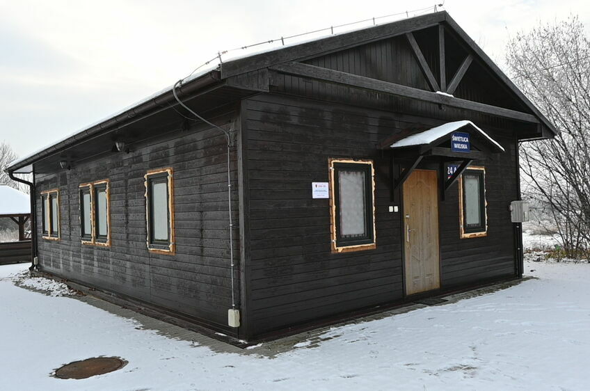 Drewniany budynek przypominający stację kolejową z tabliczkiem "Biletów" pod śniegiem, z drzewem bez liści i śladami na ziemi.