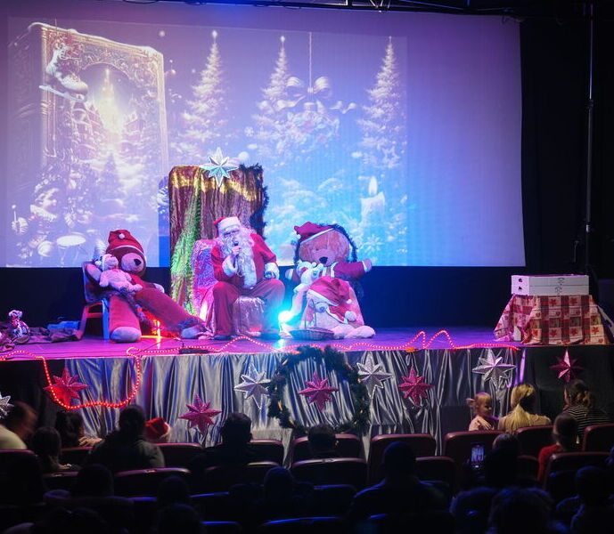 Scena teatralna z dwoma osobami w kostiumach maskotek siedzącymi po obu stronach tronu, na tle oświetlonego na fioletowo zasłania.