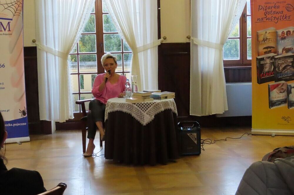 Pisarka Izabella Frączyk siedząc przy okrągłym stole z mikrofonem w ręce odpowiada na pytania widzów