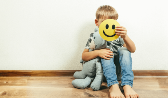 Dziecko siedzące przy ścianie z misiem, trzymające emotikonę uśmiechniętą przed twarzą