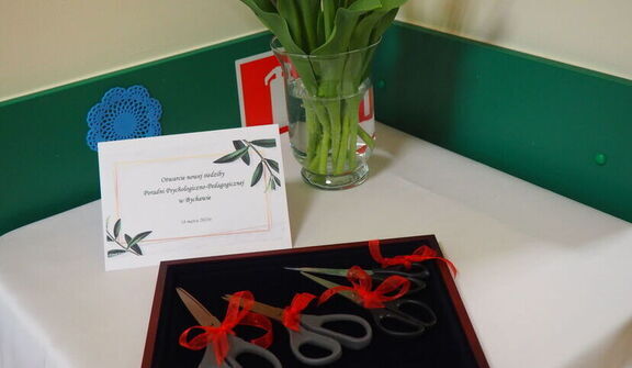 Stół przyozdobiony na uroczystość z ciętymi kwiatami w szklanym wazonie, kartką z życzeniami oraz oprawionymi nożycami z czerwoną wstążką.
