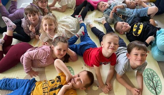 Grupa uśmiechniętych dzieci leżących na podłodze w sali zabaw, układających się w kształt gwiazdy, noszących kolorowe ubrania.