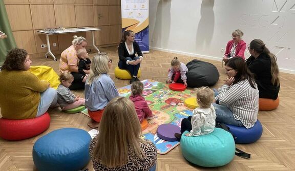 Dorośli i małe dzieci siedzą na kolorowych poduszkach na drewnianej podłodze. W środku znajduje się kolorowa mata edukacyjna. W tle widać plakat i balony. Grupa jest zaangażowana w naukę lub zabawę.