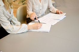 na zdjęciu znajdują się dwie kobiety dyskutujące nad leżącymi na stole teczkami z dokumentami
