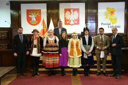 Grupa ludzi stojących w rzędzie, niektórzy ubrani w tradycyjne polskie stroje ludowe, na tle flag i herbów, w pomieszczeniu o oficjalnym charakterze.