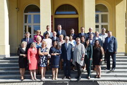 Grupa osób stoi na schodach przed wejściem do klasycznego budynku, uśmiechając się do kamery na tle żółtych kolumn i drzwi.