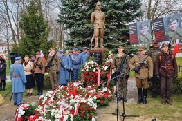 Pomnik z kwiatami na uroczystości, grupa osób w mundurach i cywilnych słucha przemówienia kobiety przy mikrofonie, w tle banery z portretami.