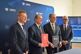 Czterech mężczyzn stoi obok siebie w pomieszczeniu z logotypami UMCS, dwóch z nich trzyma dokumenty, wszyscy uśmiechają się.