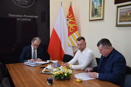 Trzej mężczyźni ubrani w garnitury siedzą przy stole z dokumentami, flagą Polski i kwiatami w tle.