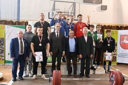 Grupa mężczyzn stoi na podium z medalem w zawodach podnoszenia ciężarów, trzymają dyplomy i trofea. W tle banery i sprzęt sportowy.