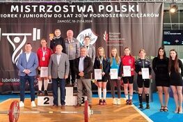 Grupa ludzi stoi na podium i obok niego podczas ceremonii wręczenia medali na mistrzostwach Polski w podnoszeniu ciężarów. Na pierwszym planie widoczny jest sprzęt sportowy.