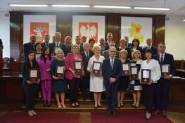 Grupa osób stoi ustawiona w kilku rzędach w eleganckim wnętrzu, trzymając certyfikaty. Na ścianie widnieją obrazy i symbole narodowe.