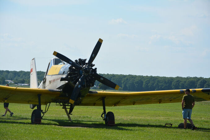 Samolot z żółtymi skrzydłami i dużym śmigłem stoi na trawiastej polanie, a mężczyzna obserwuje go z boku.