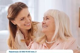 Dwie kobiety, starsza i młodsza, uśmiechają się do siebie w jasnym pomieszczeniu. Trzymają afisz z informacjami o profilaktyce raka piersi.